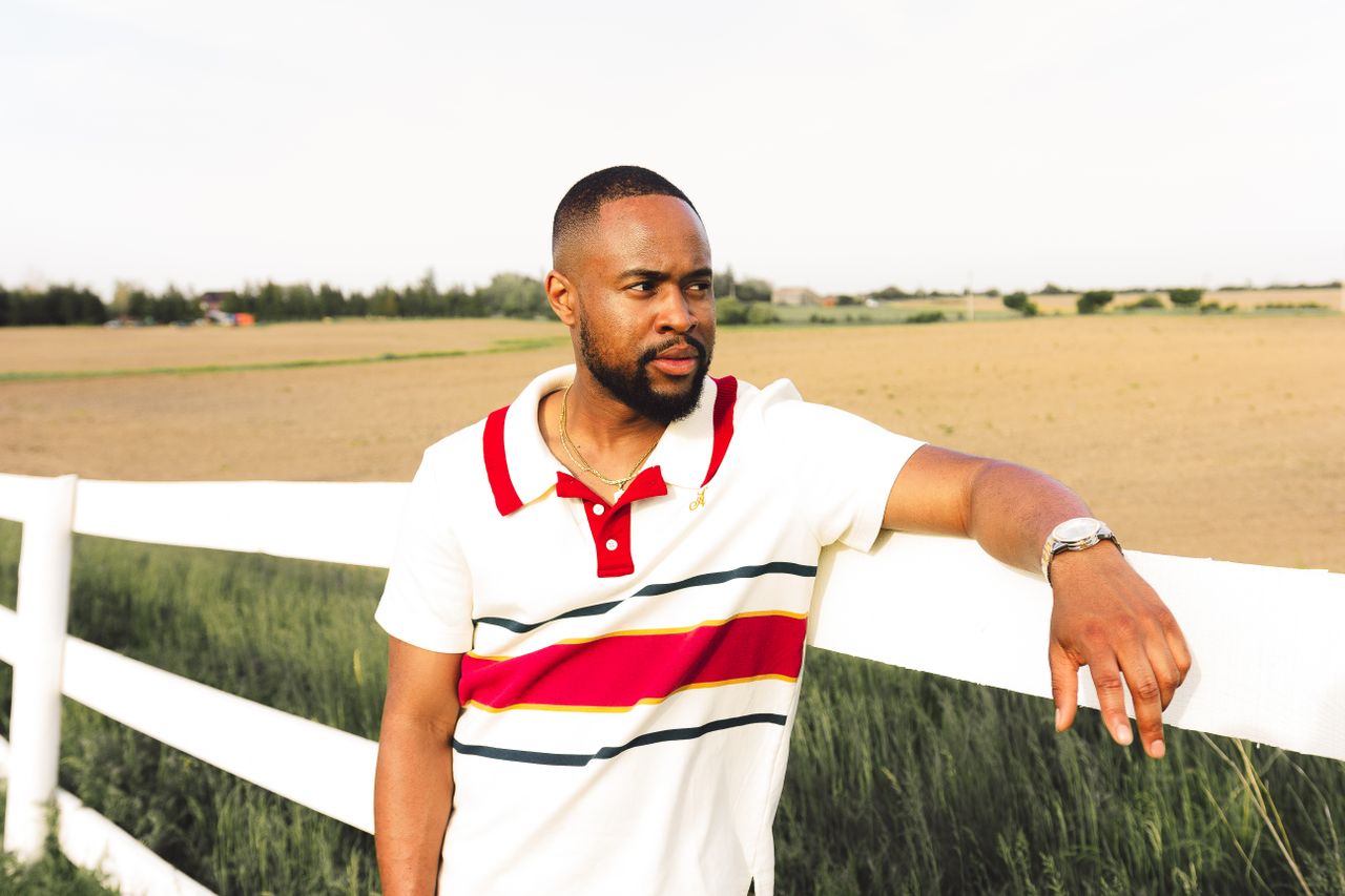 A fashionable man in casual attire leaning on a white fence in a rural setting.