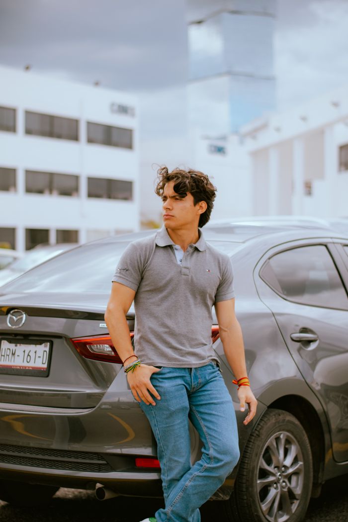 Fashionable young man in jeans and polo shirt leaning on car in cityscape setting.