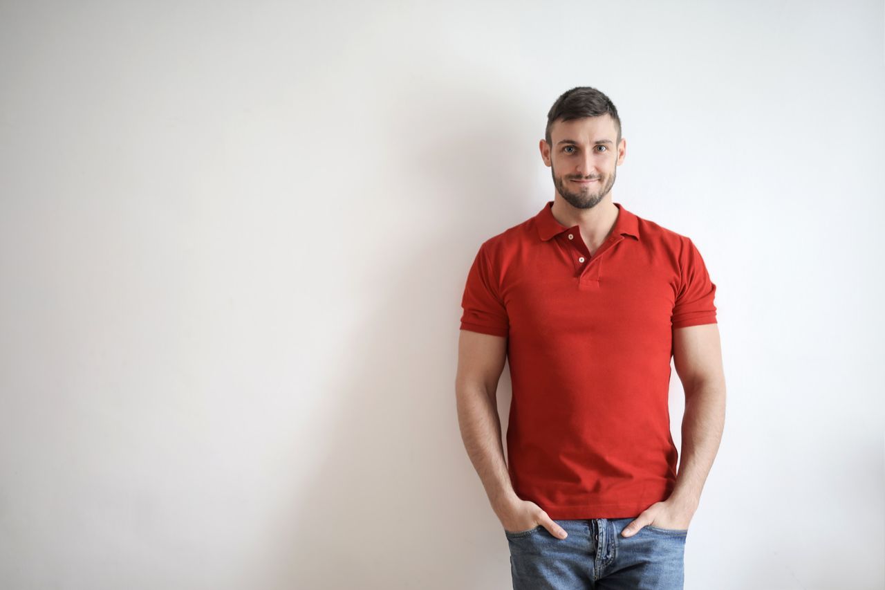 Casually dressed man in red polo stands smiling in front of a white wall.