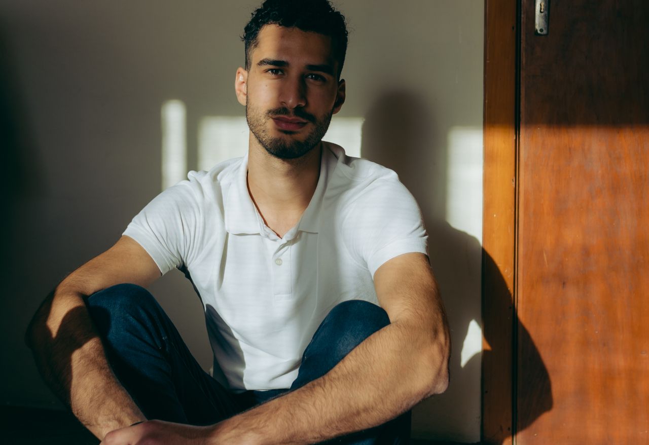 A young man in a white polo shirt sits indoors, looking at the camera with a serene expression.