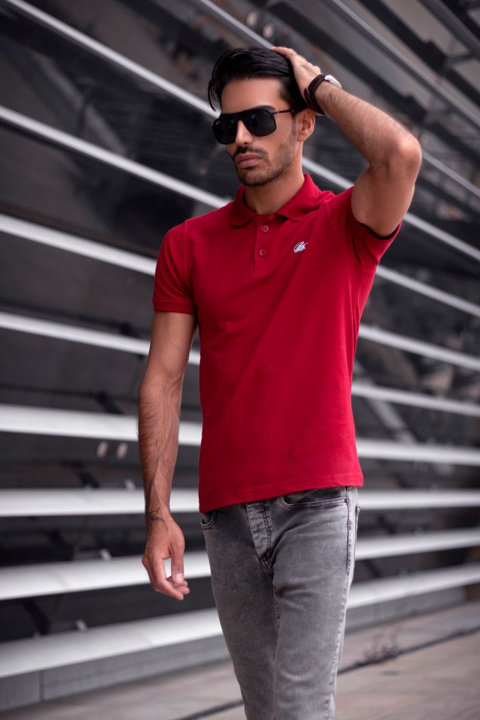 A fashionable man in a red polo shirt and sunglasses walks confidently outdoors.